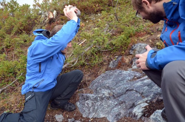 Was it like this the chert at Melsvik was quarried in the Stone Age? Archaeologist Lars Julsrud of Alta Museum applying brute force with a hammerstone (without touching the ancient surface!), his colleague Martin Hykkerud perhaps taking a more sceptic stance. Photo: Per Storemyr