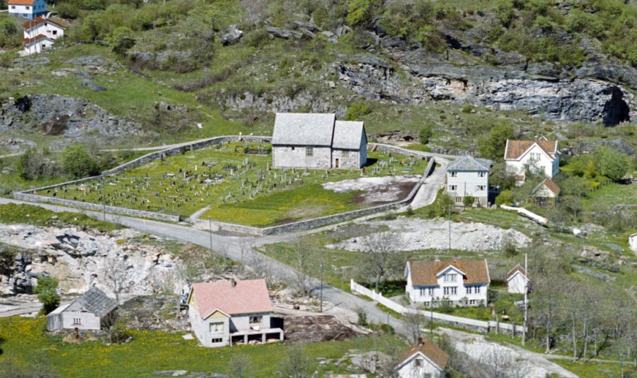 Moster gamle kirke sett rett fra sør, slik at også det store bruddet i forkant kommer med (i dag kirkegård). Bruddet i bakkant er nå Moster Amfi. Foto: Widerøes Flyveselskap 1962 (nb.no)
